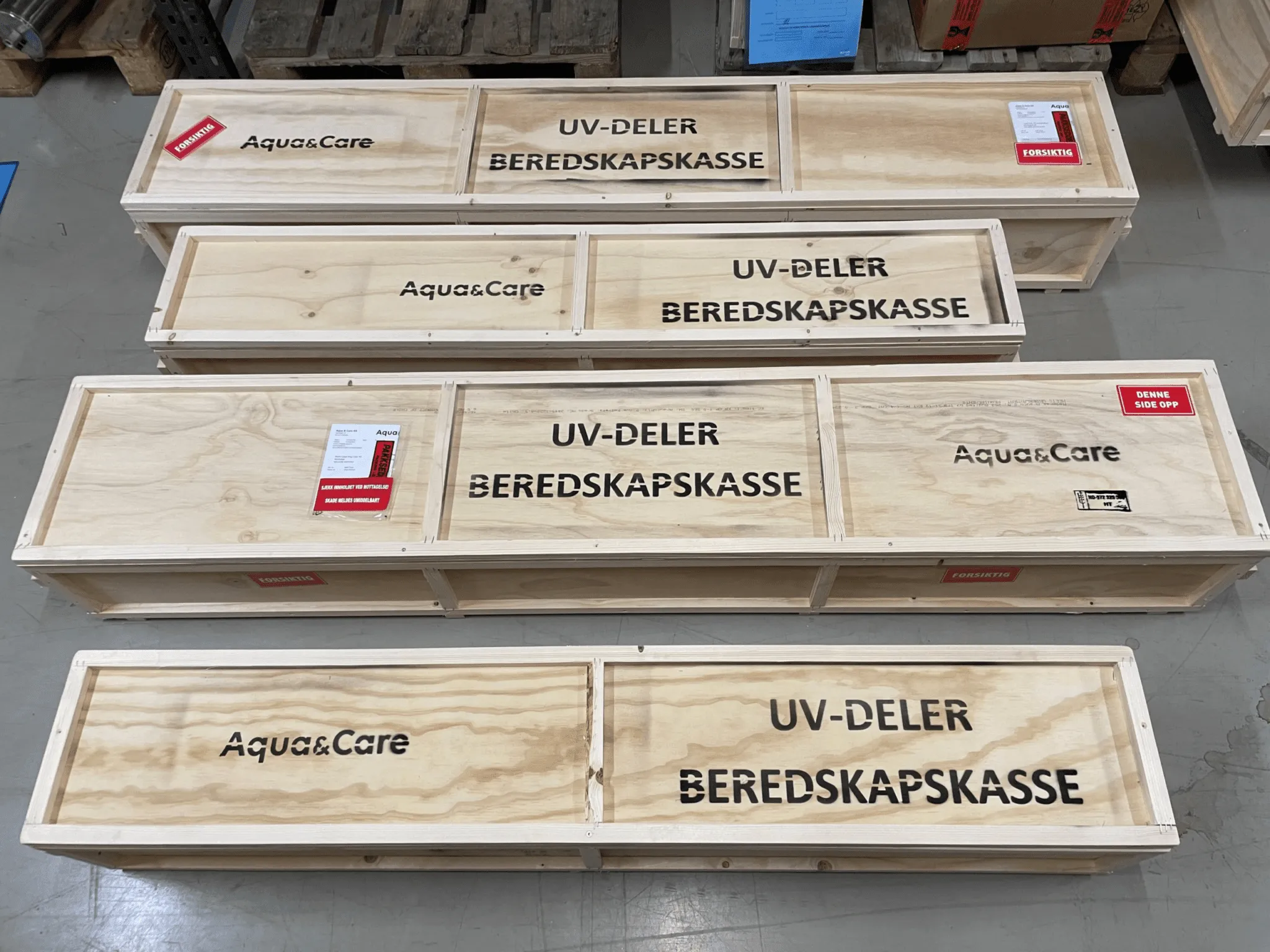 Flera beredskapslådor för UV-delar från Aqua & Care står uppradade på ett lagergolv, märkta med texten 'UV-DELAR BEREDSKAPSKASSE' och försiktighetsetiketter.