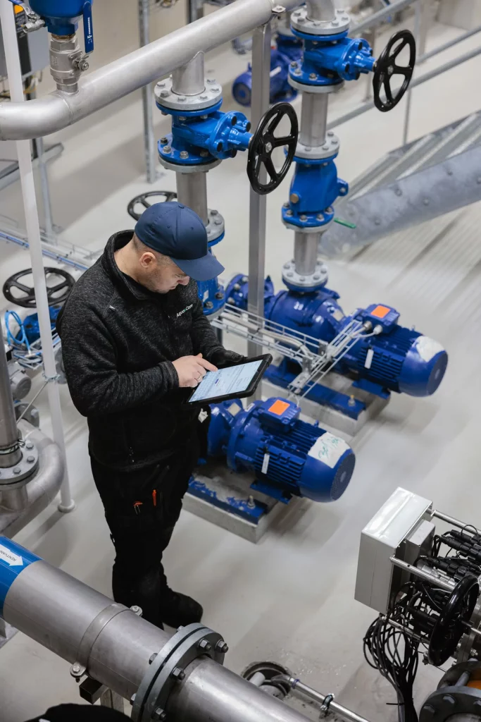 Servicetekniker från Aqua & Care utför digital kontroll av UV-anläggningens driftdata i tekniskt utrymme med pumpar och rörsystem.